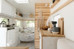Interior of a cozy camper van with a minimalist design, featuring a small kitchen area with wooden countertops and utensils, a bed with white linens in the back, and natural light streaming through the windows.