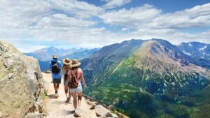Three hikers walk along a narrow mountain trail with backpacks and hats, overlooking a vast landscape of forested valleys and towering mountain peaks under a partly cloudy sky.