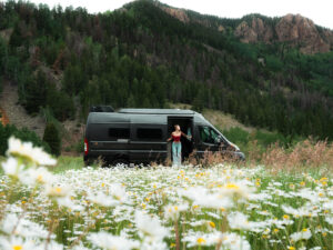 A woman stands at the open door of a black camper van parked in a field of white daisies, surrounded by lush green forest and tall rocky mountains in the background.