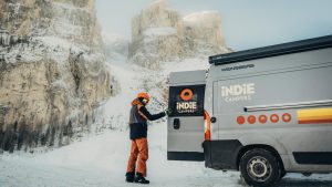 A motorhome parked beside a snow covered mountain road at sunrise, with visible snow chains on its tires and light snow falling, cozy yet adventurous moo