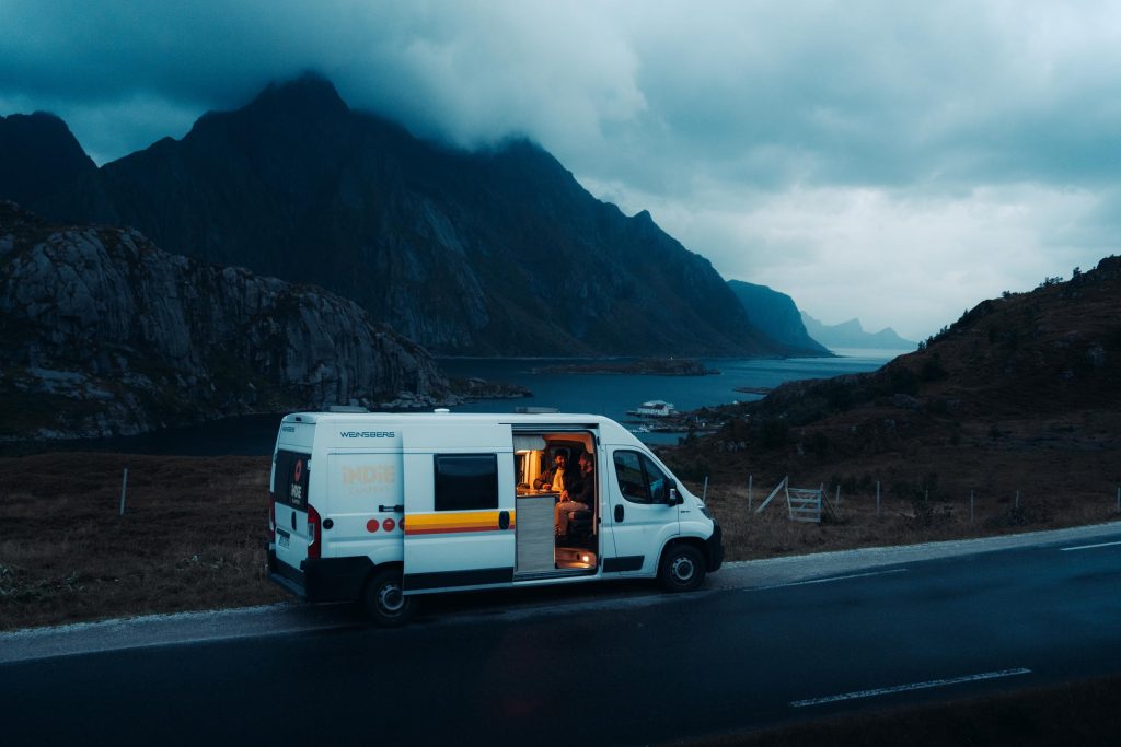 Campervan parked by a coastal road in a dramatic mountain landscape, with a person sitting inside under warm light.