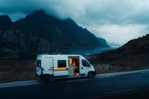 Campervan parked by a coastal road in a dramatic mountain landscape, with a person sitting inside under warm light.