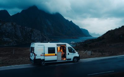 Campervan parked by a coastal road in a dramatic mountain landscape, with a person sitting inside under warm light.