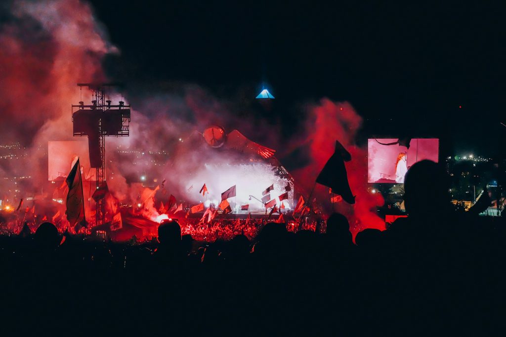 Glastonbury Festival crowd waving flags in red flare smoke in front of a stage at night
